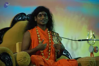 A photograph of a man seated in an ornate dark-wood and gold-upholstered throne-style chair, speaking into a microphone on a stand. He wears an orange saffron robe, a long gold bead necklace, and a red bindi on his forehead, with long swept-back dark hair. His hands are raised in gesture as he talks. Behind him is a colourful blurred backdrop of blue, yellow and orange tones with a faint trident motif, and to the right a silver trident and small drum stand beside the mic. The appearance with long hair, saffron clothing and ceremonial gold beads is a probable Swamiji (probable) on the dais giving a talk.