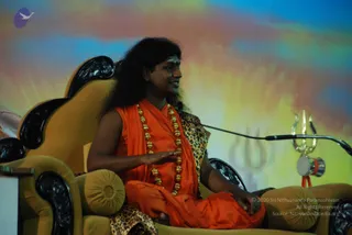 A photograph of a long-haired man in orange saffron robes seated on an ornate gold throne with carved black wooden armrests, speaking into a clip microphone on a stand while gesturing with his right hand. He wears a long string of large gold beads around his neck and a leopard-print shawl over his shoulder. Beside him on the right sits a golden trident with a small drum hung below it. The backdrop is a colourful painted sky of blue, yellow and orange with a faint trident motif. Swamiji (probable): the long swept-back hair, saffron robes, ornate gold beads and trident setting match the cues for the page's subject; he is seated speaking as a teacher on stage.