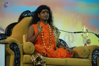 A seated man on an ornate gold-upholstered throne with carved black wooden arms addresses a satsang. He wears a sleeveless saffron-orange robe, a leopard-print shawl draped over one shoulder, a heavy gold bead necklace and a red bindi. His long dark hair is swept back, and his right hand is raised in a blessing gesture. A microphone stand and a gold trident with a small drum stand at right, and a painted sunrise-coloured backdrop with faint trident imagery fills the background.