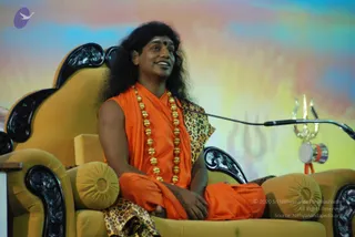 A photograph of a long-haired man seated on an ornate yellow throne-style chair with black carved armrests, speaking into a microphone on a stand. He wears an orange sleeveless robe over a leopard-print shawl and a long gold bead necklace, with a small red dot on his forehead. A golden trident and a small drum hang from the microphone stand. Behind him is a soft painted backdrop of blue, pink and yellow with a faint trident motif.
