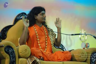 A seated man in saffron-orange robes sits cross-legged on an ornate golden-yellow throne with dark carved wooden arms and a leopard-print cloth draped over the backrest. He wears a long, thick gold bead necklace and has long, dark, swept-back hair and a small red bindi on his forehead. Both hands are raised palms-forward in a teaching gesture, and a microphone stand is positioned in front of him. To the right, a brass trident stands beside a small drum with red and orange trim. The backdrop is a soft blue, pink and yellow gradient.