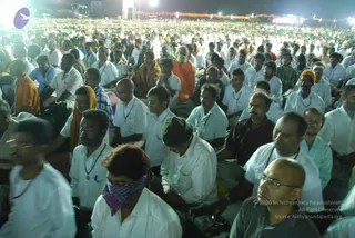 Night open-air satsang with a very large seated crowd filling the frame, most people in white shirts and some wearing lanyards, many with eyes closed in meditation. Floodlights and a row of strung lights glow in the background. In the middle distance a bearded man in an orange-saffron robe sits among the crowd, and an older bald-headed man wearing dark glasses sits in the foreground. A few men wear orange, yellow and blue cloths, and one wears a pink-and-purple checked cloth over the face. The crowd is dense across the whole frame.