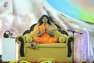 A seated man in orange robes sits cross-legged on an ornate gold throne with black carved armrests and leopard-print cushions, palms pressed together in greeting. He wears a long multi-strand gold-beaded necklace and a red bindi, with long swept-back dark hair. A microphone stand is at his right, and a small table at left holds a bottle and a brass bowl. Marigold and yellow flower garlands line the stage front, and a painted mountain backdrop with rainbow sky fills the background. Swamiji (probable): long hair, saffron robes and ornate gold necklace, seated as the speaker on the throne.