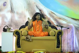 A photograph of a seated man on a gold upholstered throne with carved black wooden armrests and backrest, on a stage before a microphone on a stand. He wears a saffron-orange robe, a leopard-print shawl draped over his shoulders and lap, a long strand of gold-coloured beads, and a red bindi on his forehead, with long dark hair. He sits cross-legged with both palms raised outward in a gesture of address. Behind him is a large painted backdrop of a snowy mountain under a blue and rainbow sky. Marigold garlands line the stage edge, and a brass vessel and water bottle sit on a white side table at left.