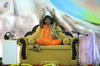 A photograph of a seated figure on a mustard-yellow armchair with black carved wooden arm panels and two round yellow cushions, on a stage with a mountain-like backdrop painted in pink, white and blue. The person wears an orange saffron robe, a leopard-print shawl draped over the shoulders and lap, a long gold-bead necklace, and a red tilak on the forehead, with long dark hair swept back; both arms are raised with palms open. Swamiji (probable): the candidate shows long swept-back hair, saffron clothing, a beaded ceremonial necklace and a shawl, consistent with the page's attribution, seated in a speaker's position. A microphone on a stand stands at the chair and a water bottle sits on a white side table.