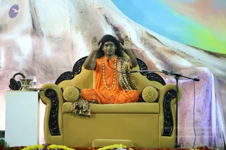 A seated man in saffron-orange robes and a leopard-print shawl sits cross-legged on an ornate gold-upholstered throne with black carved wooden arms and gold cushions. He wears a long gold bead necklace and a red forehead mark, with long dark hair, and raises both hands palms outward beside his head in a blessing gesture. A microphone on a stand is at his side. Behind him is a large painted backdrop of a snowy mountain under a blue-to-green sky, and marigold and yellow flowers line the stage edge. A white side table holds a water bottle and a small brass bowl. Probable Swamiji is present, identifiable by the saffron robes, leopard shawl, long hair, gold bead necklace and throne seating.