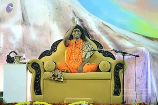 A man with long dark hair, wearing an orange robe over a leopard-print shawl and a long gold-beaded necklace, sits cross-legged on an ornate gold armchair with black carved armrests and a gold cushion. He raises his right hand in a wave toward the audience. A microphone on a stand stands beside him, and a white table with a water bottle and a small brass vessel sits at left. Marigold and yellow flower garlands line the stage front, and a painted mountain backdrop with a rainbow sky fills the background. The man's appearance is consistent with the a person cues of long swept hair, saffron clothing and ornate gold necklace, seated in a satsang setting.
