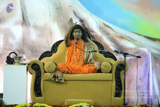 A seated man in orange robes sits cross-legged on an ornate gold upholstered throne with black carved wooden armrests and a leopard-print shawl draped over his shoulders and lap. He wears a red bindi, long dark hair, and a necklace of gold-toned beads, and raises one hand in blessing toward the audience. A microphone on a stand stands beside him. Behind the throne, a large painted backdrop of a snowy mountain under a pale sky serves as the stage set. Marigold and flower garlands are arranged at the base of the stage, and a small white table holds a water bottle and a brass vessel. The image is a photograph of a stage event. Swamiji (probable): the figure has long swept-back hair, saffron robes, a rudraksha-style gold necklace and a leopard shawl, seated on a throne, identified by these cues.