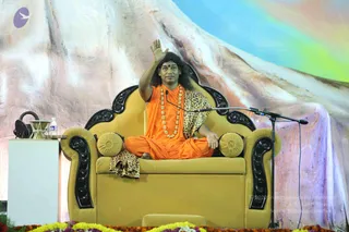 A man with long, swept-back dark hair sits cross-legged on an ornate golden-yellow throne with black carved armrests and yellow cushions on a stage. He wears a saffron-orange robe, a leopard-print wrap over his shoulder and lap, a long rudraksha-style mala of beads around his neck, and a red bindi on his forehead. He raises his right hand in blessing while a microphone stands beside him. Behind him is a large painted backdrop of a snow-capped mountain under a blue and rainbow sky, with yellow and orange flower garlands along the front of the stage, a water bottle and a small table at left. Probable Swamiji (in person), seated as a speaker during a satsang-style talk, with cues of swept-back long hair, saffron clothing and a rudraksha mala.