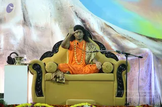 A seated figure in orange robes and a leopard-print shawl sits cross-legged on a large gold upholstered throne with black carved armrests and gold cushions. He wears a long gold-bead necklace and a red bindi, with long dark hair, and raises one hand in a blessing gesture. A microphone on a stand stands beside the throne, a water bottle and glass sit on a white table at left, and marigold and yellow flower garlands line the stage front. The backdrop is a painted mountain scene with a rainbow sky.