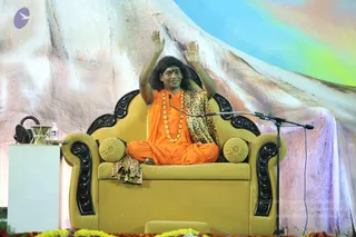 A seated man in an orange robe and leopard-print shawl raises both hands on a gold upholstered throne with carved black armrests and yellow round cushions. He wears a multi-strand gold necklace and a red bindi, with long dark hair swept back. A microphone on a stand stands beside him, and a water bottle and small table sit at left. Behind is a large painted mountain backdrop with rainbow sky, and marigold flowers line the stage front. One person is visible.