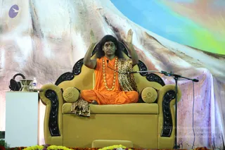 A seated man in an orange saffron robe and a leopard-print shawl sits cross-legged on a gold-upholstered sofa-style throne with black carved armrests and gold cushions. He wears a long gold-bead necklace and a red tilak on his forehead, with long swept-back dark hair, and raises both hands palms outward in a blessing gesture. A microphone stand stands before him, a glass of water and a bottle sit on a white side table to the left, and marigold flowers line the stage edge. Behind him a large painted backdrop shows a snowy mountain under a blue and green sky. The orange robe, leopard shawl and gold necklace are visible cues for a person, seated in the speaker's role during a satsang-style talk.