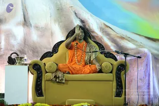 A photograph of a man seated cross-legged on an ornate olive-gold throne with black carved wooden arms and two round gold cushions, raising one open right palm in a blessing gesture. He wears a saffron-orange robe, a long beaded gold-toned necklace, and a leopard-print shawl draped over his shoulder and lap. A microphone on a stand is at his side, a glass of water sits on a white pedestal to the left, and marigold-type flowers line the stage edge. Behind him is a large painted backdrop of a snowy white mountain under a blue and rainbow sky. Swamiji (probable) is seated in the centre, identified by the saffron robe, long swept-back hair, ornate gold necklace and throne seating.