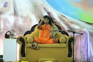 A photograph of a long-haired man in saffron-orange robes seated cross-legged on an ornate mustard-yellow throne with black carved armrests and yellow round cushions. He wears a long beaded necklace of gold-toned beads, a leopard-print shawl draped over one shoulder, and a red tilak on his forehead. He raises his right hand in a blessing or teaching gesture toward the audience. A microphone stand is in front of him, a white side table holds a water bottle and a brass vessel, and marigold flowers line the stage edge. The backdrop is a painted mountain scene with pastel blues and pinks.