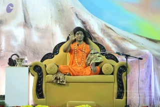 A photograph of a man seated cross-legged on an ornate gold throne with black carved armrests and gold cushions. He wears an orange saffron robe and a long gold-beaded necklace, with a leopard-print shawl draped over his shoulder and lap. His long dark hair is swept back, and his right hand is raised in a blessing gesture. A microphone on a stand stands beside him, and a small table with a glass and water bottle sits at left. Behind him is a large painted mountain backdrop with blue and pastel tones, and marigold flowers line the foreground. a person is seated in the speaker's position, giving a satsang discourse.