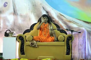 A seated man in an orange-saffron robe and gold-bead necklace sits cross-legged on an ornate olive-gold throne with black carved armrests and gold cushions, raising one hand in a blessing gesture. He has long dark hair, a red bindi and a leopard-print shawl draped over the chair. A microphone on a stand is at his side, a water bottle and glass sit on a white table to the left, and marigold garlands line the stage front. Behind him a large painted backdrop of snowy mountain peaks under a pale blue and rainbow sky. Appearance-based cues (long swept hair, saffron robe, gold necklace, throne, orange clothing) match the a person candidate, presented as a speaker at a satsang stage.