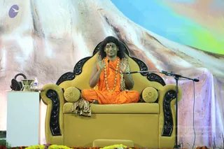 A photograph of a man seated cross-legged on an ornate gold-yellow throne with black carved wooden armrests and backrest, flanked by yellow round cushions and a leopard-print cloth draped over the seat. He wears a saffron-orange robe, a long gold bead necklace, and has long dark swept-back hair with a red bindi, raising both palms in a gesture of address while speaking into a microphone on a stand. Marigold flowers line the stage edge in front of a large painted backdrop of snowy mountains under a rainbow sky. A white side table holds a water bottle and a glass. Swamiji (probable): long swept-back hair, saffron robe and gold bead necklace, seated on the throne as speaker.
