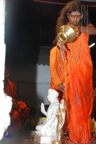 A standing man in orange saffron robes with long, swept-back curly hair and a red tilak on his forehead pours water from a brass kalasha onto a white marble seated deity murti. A gold-beaded mala hangs around his neck. Behind him, a second figure in orange also stands, and another orange-robed devotee sits in the left background. A brass-coloured small deity statue stands beside the white figure. The scene suggests an abhishekam in a dim temple hall.