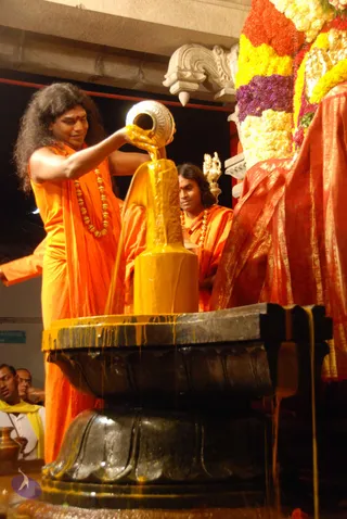 A photograph of a Shiva linga abhishekam at night. A man with long, curly, dark hair and a red bindi, wearing saffron-orange robes and a gold bead mala, pours yellow liquid from a silver vessel over a black stone linga set in a black yoni-pitha base. Behind him a second man in orange robes and a gold bead necklace stands watching, and a silver ritual object is visible. To the right, a deity murti is draped in red and gold silk and decorated with yellow and red marigold garlands. Yellow liquid streams down the linga and pooling base. Two men in white and yellow attire stand at the left edge.