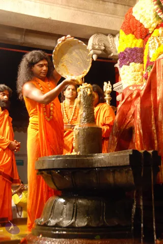 A long-haired man in saffron-orange robes and a rudraksha-style bead necklace pours a liquid from a large brass vessel over a dark black stone Shivalinga on a circular yoni-pitha base during abhishekam. Behind him, several men in orange robes stand, one with a bead mala and another with hands folded. At right, a deity murti is draped in red and gold cloth and adorned with a large garland of yellow, red and purple flowers. Liquid runs down the lingam and pools on the base, with a brass lamp visible in the background.