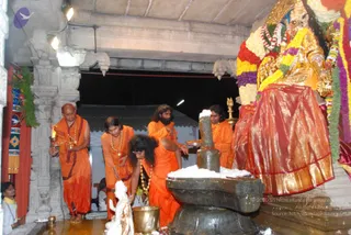 Photograph inside a temple sanctum during a Shiva linga abhishekam. A dark stone linga on a yoni pedestal is being bathed, with white milk or sacred substance heaped around its base. Several priests in saffron-orange robes stand and kneel around it; a man on the left holds a lit oil lamp and a brass plate, a man in the centre pours from a vessel, and a bearded man in orange holds a small tray. A brass kalasha stands in front. Behind them a gold-and-red deity murti is adorned with marigold, yellow and red flower garlands and a gold crown. Flowers, marigold and greenery decorate the pillars and archways. No individual is identified from appearance; the priests are described by role.