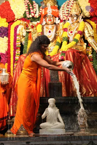 A person in an orange saffron garment with long, swept-back dark hair pours water from a silver vessel over a white marble seated figure, in a temple abhishekam. Behind, golden deity murti stand adorned with red and gold garlands, wearing red silk drapery and jewelled necklaces. A dark black linga stands on a pedestal beside them, and a metal pyramid-shaped object sits at the lower left. Another person in orange stands at the left edge holding a silver pot. Water streams down onto the marble figure and the stone base.