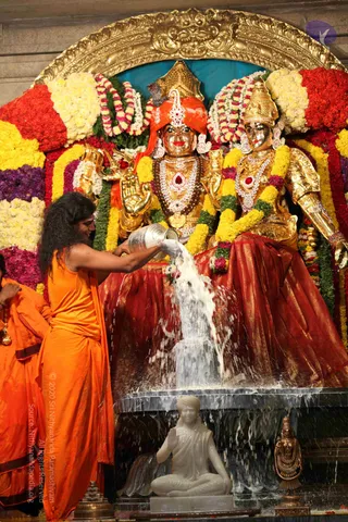 A photograph of a temple abhishekam in progress before a gilded deity pair decorated with marigold and red flower garlands. A standing figure with long, dark, wavy hair wears a saffron-orange sleeveless garment and pours milky liquid from a steel vessel over the central metal murti and its base. The deities, one with a red turban and crown and the other in a gold crown with a red skirt, are adorned with jewelled necklaces and rudraksha beads. In the foreground, a small white marble seated figure with raised hand sits below the platform, and another orange-robed person stands at the left edge. Liquid splashes across the marble base.