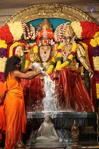 A photograph of an abhishekam at a temple shrine. A figure with long dark hair and an orange robe pours milk from a silver vessel over a golden deity murti of a seated divine couple. The murti wears a gold crown, red and white jewelled necklaces, and a red silk skirt, and is framed by garlands of yellow, red and white flowers. A small white marble seated figure sits on the platform below, and a bronze lamp stand is at the lower right. Milk cascades over the stone base in a white stream. Only the orange-robed figure pouring is a physical person visible, so no other people count. The pouring figure is a priest performing the ritual, not an identified person.