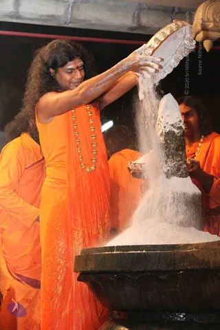A man with long, curly, swept-back dark hair and a red tilak on his forehead pours white powder from a silver plate onto a black stone Shivalinga resting in a dark basin. He wears a saffron-orange garment draped over one shoulder and a heavy gold bead necklace. Behind him, another saffron-clad man with long hair and a gold necklace holds a small silver vessel near the linga. Another orange-robed figure stands partly visible at left. The powder covers the linga and basin, indicating an abhishekam ritual.