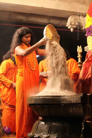 A long-haired man in saffron-orange robes and a rudraksha mala pours a stream of white powder from a golden plate onto a black stone linga-shaped base during an abhishekam. A red tilak marks his forehead. Two other saffron-clad attendants stand behind him, and a brass lamp and a garland of yellow and red flowers appear at the right edge.