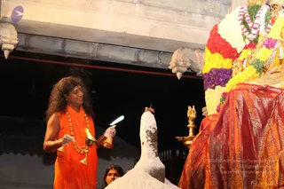A temple scene at night in which a man in an orange-saffron robe with long, swept-back curly hair and a red tilak on his forehead stands at left holding a brass oil lamp with a flame in one hand and a brass bell in the other, both hands partly covered in white. He wears a beaded rudraksha-style mala with a ceremonial necklace. In front of him is a white powder-covered linga with a red flower on top, being prepared for abhishekam. Behind and to the right, a large deity murti is draped in red and gold silk and decorated with garlands of yellow, orange, red and purple flowers. A brass oil lamp stands nearby and a woman in a dark outfit is partly visible at the bottom edge.