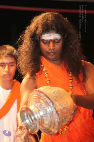 A man with long, voluminous curly dark hair and a white horizontal tilak on his forehead, wearing an orange saffron wrap and multiple gold ornate beaded necklaces, holds a large ornate silver kalasha vessel with both hands and pours water from it during an abhishekam. He looks downward in concentration. Behind him to the left, a younger man in a white garment with an orange sash and a red bindi stands in the dark background.