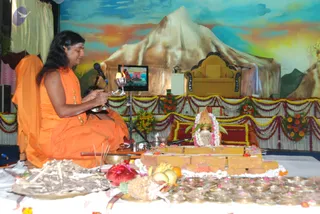 A photograph of a ritual scene with a man in long swept-back dark hair seated at left, wearing an orange-saffron robe and gold-toned necklaces, holding a silver vessel and a small object while seated on a low wooden platform in front of a microphone stand. Behind him a small monitor shows a temple image. At right a deity murti or linga stands on a yellow-brown brick-like altar garlanded with white and pink flowers, with a bowl of fruit, bananas and a pineapple, and marigold garlands. A painted backdrop shows a snowy mountain and golden sky, with an empty golden throne. Dried wood sticks lie in the foreground.
