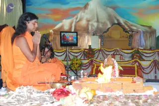 A homa fire ceremony on a stage decorated with marigold and flower garlands. A long-haired figure in orange robes sits cross-legged at left with palms pressed together in namaste, a small ornament at the neck, while a second person in orange sits behind, holding a long ceremonial stick. In front, a fire burns on a raised altar of wooden pieces, with fruits, flowers, a brass bowl and incense sticks around it. Behind, a painted mountain backdrop sets off an ornate gold throne-like seat, and a small television screen shows a live feed of the ceremony. a person: the long-haired figure in orange with swept-back hair and ceremonial jewellery, seated in a priestly role at the homa.