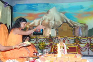 Photo of a homa fire ceremony on a stage backdrop painted with a snow-capped mountain under a sky of blue, orange and yellow. A man with long dark hair and white ash markings on his forehead wears an orange robe and holds a silver tray of flowers and food while extending his other hand over a fire pit. Behind him, a second orange-robed man sits in a similar pose. The fire rises from a brick-lined altar decorated with flowers. An ornate golden throne stands empty at the back of the stage, framed by marigold and rose garlands and yellow-and-purple festoons. A small screen and microphones are visible on the stage.