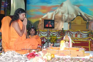 A photograph of a fire ritual on a stage. A long-haired figure in orange robes sits cross-legged at left with palms pressed together in namaskar, a microphone before him. Beside him a younger person in orange with a beaded necklace sits behind a brass bowl. At centre, a flame rises from a brick homa pit decorated with flowers, fruit and a kalasha with a coconut and mango leaves. Wooden sticks and offerings lie in the foreground. Behind them a painted backdrop shows a snow-capped mountain under a colourful sky, marigold garlands drape the stage, and an empty ornate golden throne stands at right. A television in the background shows a figure in orange speaking, consistent with a live broadcast of a homa ritual.