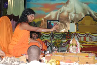 A photograph of a homa-style ritual on a stage. A man with long swept-back dark hair, wearing an orange saffron garment, a rudraksha-style gold bead necklace and a sandalwood-marked forehead, sits on the left and holds a round brass plate over an offering area. Behind him, a second orange-clad person sits with a shaved head. In the foreground, a shaved-head devotee in white sits before a low brick fire pit strewn with bananas, oranges, marigolds and wood sticks. Behind the altar stands a painted mountain backdrop, a golden throne draped with flower garlands, a framed picture and a decorated Shiva linga surrounded by flower garlands and a kalasha.