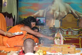 A photograph of a homa ritual. A long-haired man in orange robes with a gold necklace leans over a brick fire pit, pouring offerings into rising smoke, while a second orange-robed priest sits beside him. A shaved-headed devotee in the foreground is seated before the altar. Offerings of fruit, flowers, coconut and a silver tray of white material sit on the platform, next to a decorated kalasha wrapped in a garland. Behind them hangs a painted mountain backdrop, a gold throne, and draped festoons of marigolds and gold cloth. The man with the swept-back long hair and gold necklace has distinctive ceremonial cues, so he is a probable Swamiji candidate officiating the homa.