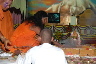 A photograph of a ritual at a decorated altar. A figure in orange robes with long, swept-back dark hair and gold ornaments on the arm sits cross-legged at the right, leaning over a tiered yellow-ochre platform holding a small kalasha with white flower garlands and marigold offerings. A shaved-headed person in white stands in the foreground, and an orange-robed attendant with a shaved head sits at the left near brass plates and incense sticks. Marigold and red-flower garlands drape the altar, and a screen shows a painted mountain backdrop behind. a person: the robed figure in orange with long swept-back hair and gold adornments, performing the homa/abhishekam offering.