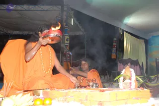 A photograph of a homa ritual at night under a tent. A man with long dark hair and a flowing orange robe with gold beaded necklaces holds a red-and-silver vessel with a flame rising from it, while another man in an orange robe and gold necklace sits behind a small offering table. Smoke fills the air. On the table are bananas, orange fruits, a metal pot, brass vessels, a white flower garland and a decorated kalasha. Blue and orange draped cloth hangs in the background. Two people are visible.
