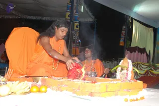 Night outdoor ritual under a tent canopy. A man with long swept-back dark hair and a tilak on his forehead, wearing orange robes, rudraksha-style beaded necklaces and gold beads, pours or offers from a red cloth-wrapped vessel over a yellow brick fire pit (homa kunda) where flames and smoke rise. A second person in orange robes and gold necklace sits beside him with a small metal kettle-like pot. Offerings of bananas, oranges and a flower-garlanded kalasha with mango leaves sit nearby, and a microphone stands in front. Decorated pink and white drapes and colourful banners frame the stage, with seated devotees visible in the background.