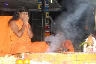 A photograph of a long-haired man in saffron-orange robes and a gold bead necklace sitting cross-legged with palms pressed together in namaskar at a ritual platform. A tilak mark is on his forehead. Behind him a second person in orange robes sits by a silver kalasha and brass vessels as smoke rises from a fire ritual. Fruit, flowers and a flower-garlanded pot sit on the yellow platform, and a microphone stands nearby under a tent with painted colourful pillars. a person: the seated man has long swept-back hair, saffron robes and a gold necklace, though the appearance is only partly clear.