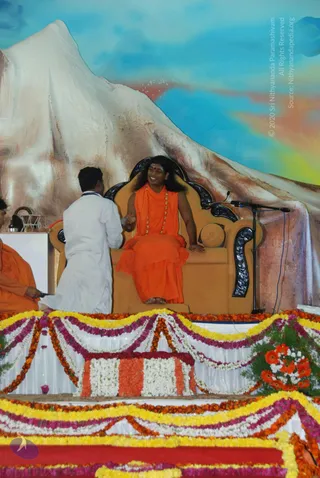 Photograph of a stage at a satsang event. A seated figure in an orange saffron robe, with long dark hair, a red tilak on the forehead and a long gold-beaded necklace, sits smiling on an ornate tan throne with black carved armrests, a microphone stand beside him. A man in a white shirt and white dhoti kneels at the left side of the throne, offering something in his hands. Another person in orange robes sits at the far left edge beside a small table with a brass vessel. Behind the stage is a painted backdrop of a snow-capped mountain under a blue sky. The stage front is decorated with layered garlands of marigolds and white and magenta flowers, with orange gerbera arrangements at the right corner.