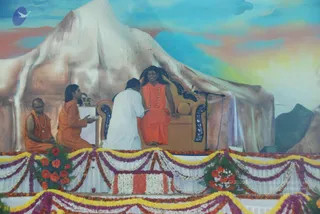 A stage decorated with marigold and flower garlands and a painted mountain backdrop with blue sky and sunset tones. A man in orange robes with a long hair and beaded necklaces sits on an ornate brown throne-like armchair beside a microphone stand. Two orange-robed men sit on the stage to the left, one with a shaved head holding a bowl and the other with long hair near a small table. A devotee in white clothes kneels in front of the throne, facing it with hands together. Flower-covered stage edges and a small floral arrangement surround the scene.