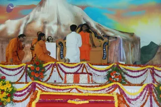 A stage decorated with marigold and flower garlands and a painted backdrop of a snow-capped mountain under a colourful sky. A long-haired figure in orange robes with a beaded necklace sits smiling on an ornate golden throne-style chair, while a younger man in a white kurta kneels before him with hands folded. Behind them, several people in orange robes sit on the stage, one with hands in prayer, and a white-draped table holds small items. Swamiji (probable) is seated on the throne in orange robes with a rudraksha-style necklace and long swept-back hair, presiding over the discourse as the kneeling devotee offers respect.