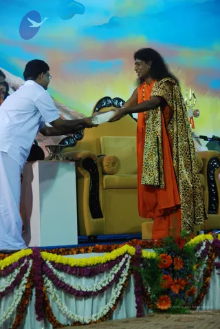 A photograph of a stage event with a bright painted sky backdrop. A man in white shirt and white dhoti, wearing glasses, leans forward and hands a folded white cloth or paper to a long-haired man in orange robes draped with a leopard-print shawl, wearing a gold necklace. The long-haired man stands beside a gold velvet throne-style chair with dark carved armrests. A trident and a small metal stand sit nearby, and the stage edge is decorated with marigold and orange flower garlands. Two people are in the scene; the orange-robed man displays distinctive long swept-back hair, saffron robes, a leopard shawl and a gold necklace, matching the Swamiji (probable) cues, while the white-clad man is a devotee presenting an offering.