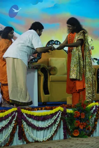 A photograph on a stage with a painted blue and yellow sky backdrop and marigold and white flower garlands draped along the front edge. A man in a white shirt and cream dhoti bends forward to hand a white envelope or card to a long-haired figure in orange robes and a leopard-print shawl, who sits in an ornate gold armchair with dark carved armrests. Behind them a second man in orange robes stands at left, and a small trident-like ornament appears at right. The seated figure has long swept-back hair, a crown-like adornment, rudraksha mala beads and a red-orange garment, with an orange-and-yellow flower arrangement on the stage in the foreground. Probable Swamiji appearance cues (long hair, saffron robes, rudraksha mala, ornate seating) for the seated figure, who is receiving the offering; the attendants are unidentified.
