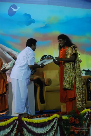 A photograph of a stage event with a painted blue sky and clouds backdrop and a floral garland-draped dais. A man in white shirt and white dhoti, bending forward, hands a white certificate or paper to a long-haired man in an orange-red robe with a leopard-print shawl and a beaded mala necklace. The man receiving the paper smiles broadly and extends both hands. Gold-upholstered armchairs stand behind them, and a partially visible person in orange clothing stands at the left edge holding a book. Marigold and orange flower garlands line the stage front. The receiving man in orange and leopard print has distinctive long swept hair and a mala, a probable match for a person, but identity is uncertain from appearance alone.