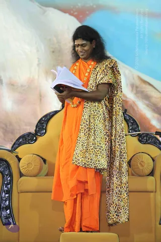 A photograph of a man standing barefoot on a raised yellow platform in front of an ornate golden throne-style chair with carved black trim and round tufted cushions. He wears an orange saffron robe with a leopard-print shawl draped over one shoulder, a gold bead necklace, and a red tilak on his forehead. He holds open a book or printed document and looks down at it, smiling. The backdrop shows a painted snowy mountain and blue sky.