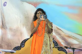 A smiling man with long dark swept-back hair and a red bindi stands on an ornate gold-and-black throne-like seat in front of a painted backdrop of snowy mountains and a blue sky. He wears an orange saffron shawl over his shoulders, a multi-strand gold bead necklace, and a leopard-print garment over one arm. He holds up a small card bearing a photograph of a person in orange, and a microphone stand is visible at the right edge. The setting resembles a stage at a public talk. Swamiji (probable) is shown in person, identified by the long swept-back hair, saffron clothing and gold necklace.